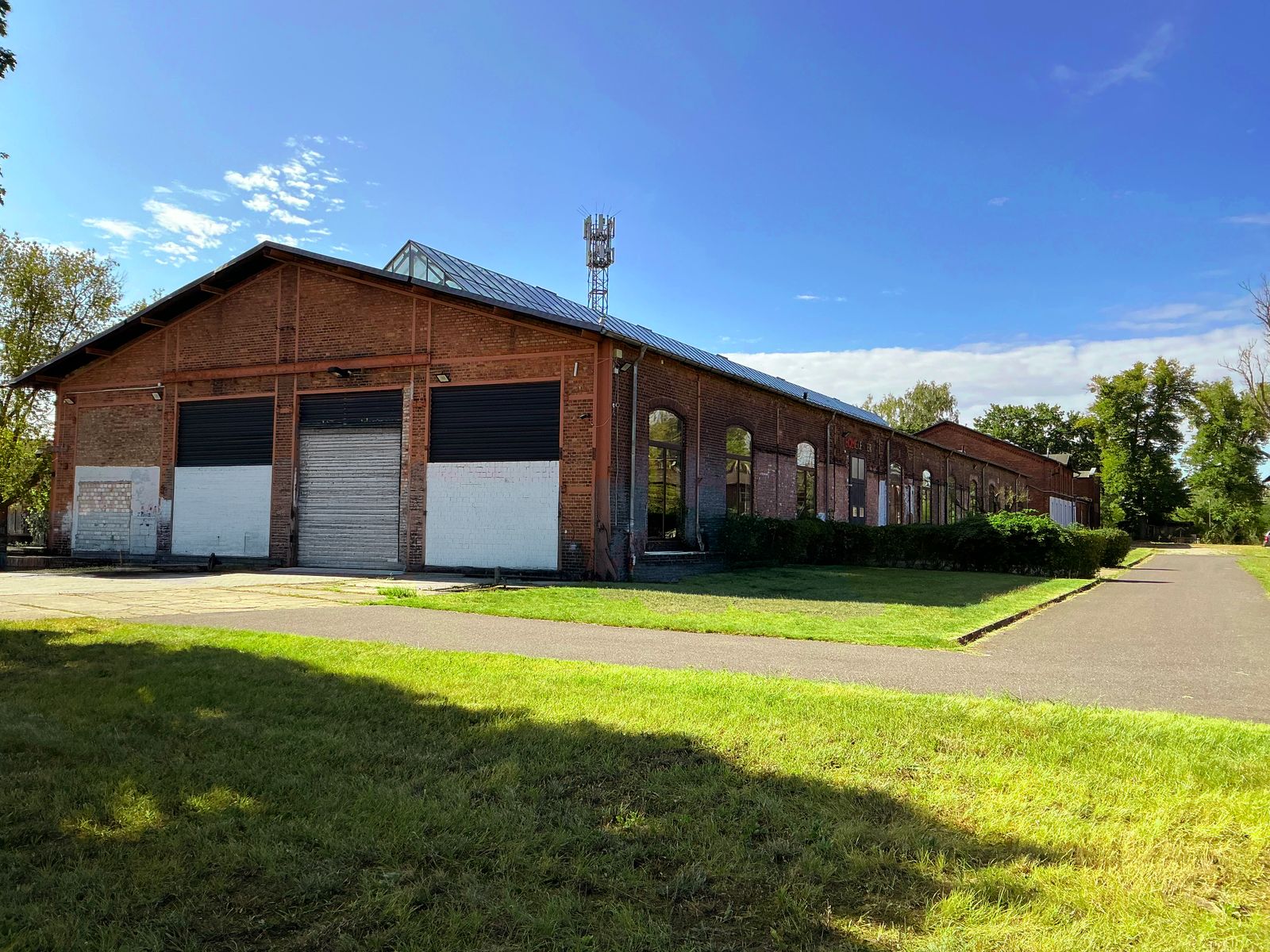 Die Backsteinhalle des Cordeshofs an einem Sommertag: Rolltore, Rasen, Zufahrt und blauer Himmel über Grunewald
